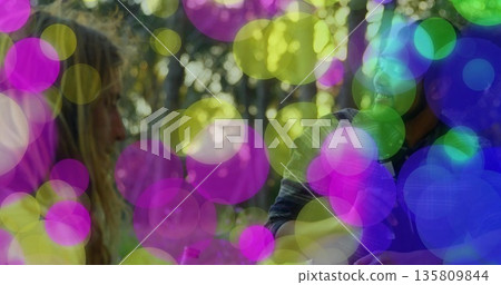 Friends standing in casual tops facing each other in wooded park, with headphones and pink bottle 135809844