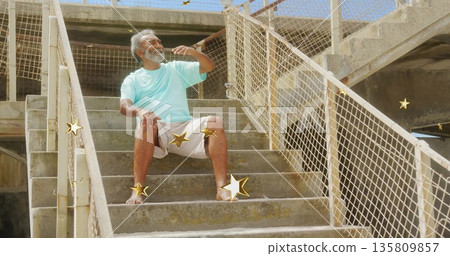 Sitting man in T-shirt making phone gesture on stairs under bridge, with mesh rails, star overlays 135809857