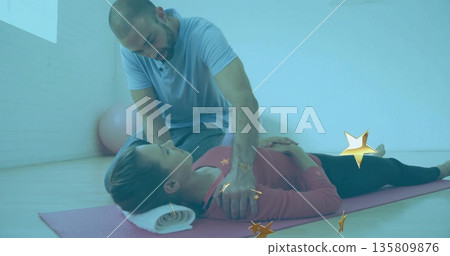 Kneeling therapist guiding client posture on pink mat in therapy studio, with rolled towel and ball 135809876