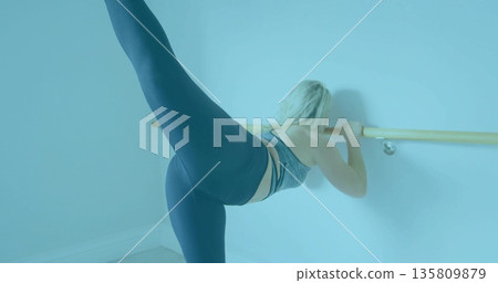Stretching woman wearing grey bra, black leggings using wooden ballet barre leg stretch in studio 135809879