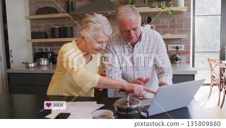Leaning senior couple working on laptop at home kitchen island with papers pen French press mug Leaning senior couple working on laptop at home kitchen island with papers pen French press mug 135809880