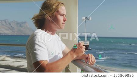 Leaning man holding glass coffee mug at metal railing on seaside deck, with white support column 135809886