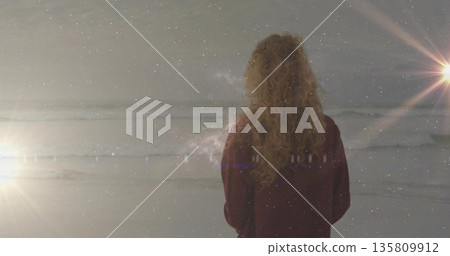 Standing woman wearing red jacket facing ocean waves on sandy beach at dusk, with lens flares 135809912