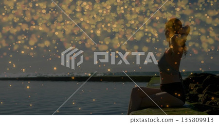 Sitting athlete wearing sportswear on rocky ledge by water at sunrise with golden orbs, copy space 135809913