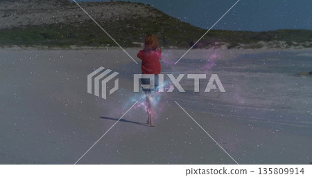 Walking barefoot woman wearing red long-sleeve top and light shorts on sandy beach, waves lapping 135809914