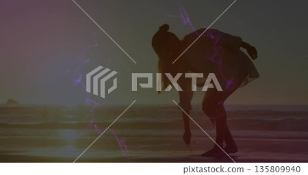 Solitary woman in tunic bending touching wet sand on beach, with waves purple streaks sun dipping 135809940