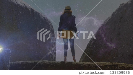 Standing explorer wearing wide-brimmed hat and dark jacket on rocky ledge at night, with starry sky 135809986
