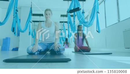 Meditating woman wearing white tank top, leggings on mat at studio, with sky blue aerial hammocks Meditating woman wearing white tank top, leggings on mat at studio, with sky blue aerial hammocks 135810008