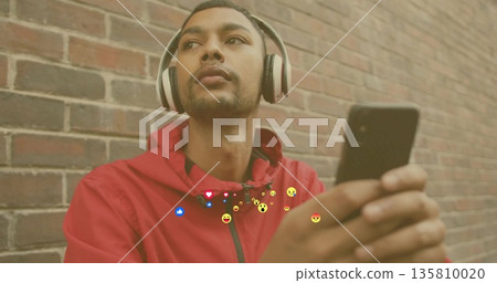 Leaning Indian man in red hoodie and headphones checking smartphone on brick wall, floating emojis 135810020