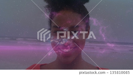 Smiling woman wearing red top standing on sandy beach edge with waves, pink-purple nebula overlay 135810085