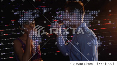 Colleagues wearing cocktail attire sipping from glasses at reception, LED world map display 135810091