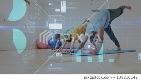 Holding downward dog pose partners practicing yoga in fitness studio, with mats, balls, data charts 135810142