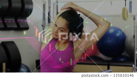 Stretching woman in magenta top tying hair at gym, with dumbbells, exercise ball, chart overlay 135810146
