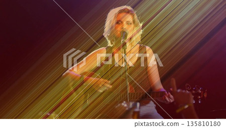 Strumming acoustic guitar, teenage musician singing into mic at club under light beams, wristband Strumming acoustic guitar, teenage musician singing into mic at club under light beams, wristband 135810180