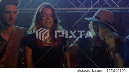 Singing bandmates on stage near metal truss, holding microphone and playing guitar under lights 135810191