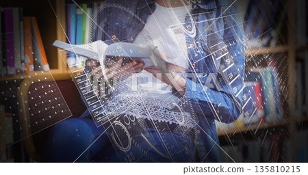 Reading teenage student holding hardback book in library seating area, with holographic charts Reading teenage student holding hardback book in library seating area, with holographic charts 135810215