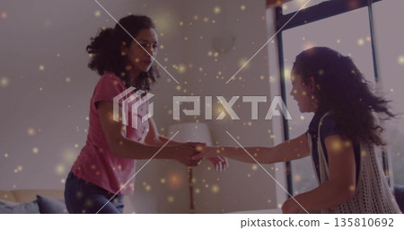Spinning mother and daughter in living room near sofa's floor lamp by window, with golden specks Spinning mother and daughter in living room near sofa's floor lamp by window, with golden specks 135810692