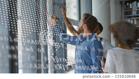 Collaborating professionals sketching diagrams on glass whiteboard in office with sticky notes Collaborating professionals sketching diagrams on glass whiteboard in office with sticky notes 135810698