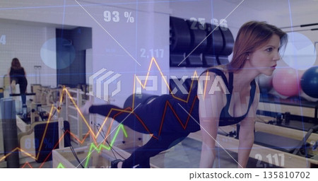 Woman in black activewear extending leg on reformer in pilates studio, with graphs, copy space Woman in black activewear extending leg on reformer in pilates studio, with graphs, copy space 135810702