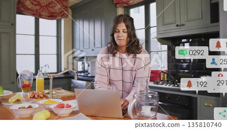 Working woman typing on laptop at home kitchen island, with blender pitcher and sliced fruit Working woman typing on laptop at home kitchen island, with blender pitcher and sliced fruit 135810740