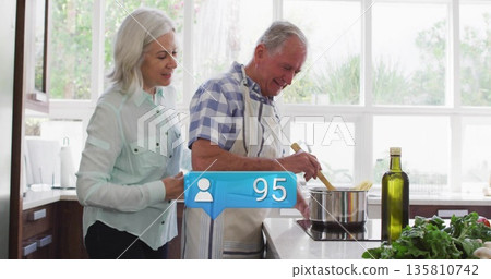 Cooking senior couple stirring sauce in saucepan at kitchen island, with olive oil and fresh greens Cooking senior couple stirring sauce in saucepan at kitchen island, with olive oil and fresh greens 135810742