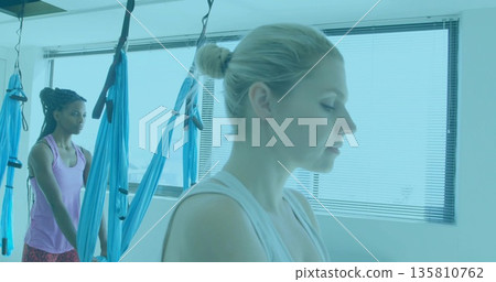 Balancing woman wearing white tank top doing aerial yoga at studio, with blue hammocks, copy space 135810762