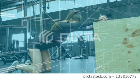 Kneeling woman stretching torso forward on padded mat in gym wearing dark top, using plyometric box 135810772