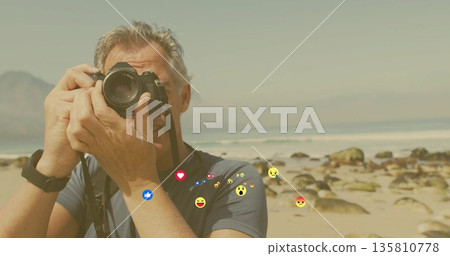 Holding DSLR camera to eye, photographer capturing ocean waves at sandy shoreline, floating emojis 135810778