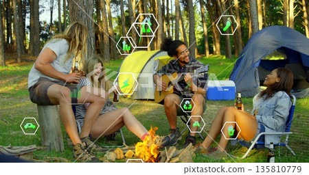 Friends playing guitar, holding beer bottles at campfire in forest clearing, with yellow or blue ten Friends playing guitar, holding beer bottles at campfire in forest clearing, with yellow or blue ten 135810779