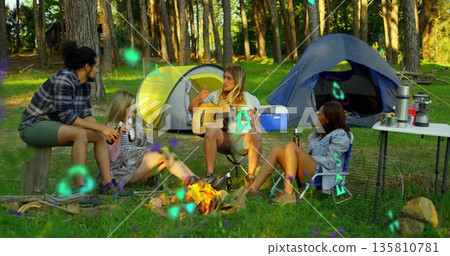Sitting around campfire four friends chatting in forest clearing, with acoustic guitar, tents Sitting around campfire four friends chatting in forest clearing, with acoustic guitar, tents 135810781