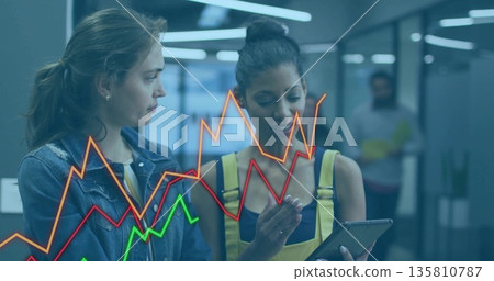 Two coworkers wearing yellow overalls studying tablet in office corridor, with chart overlay 135810787