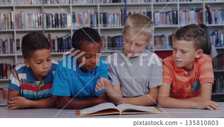 Leaning four school age boys reading open hardcover book in library reading area, with bookshelves Leaning four school age boys reading open hardcover book in library reading area, with bookshelves 135810803