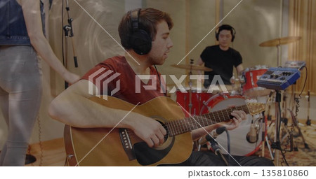 Strumming guitarist in red tee and headphones in studio, with acoustic guitar and drum kit 135810860