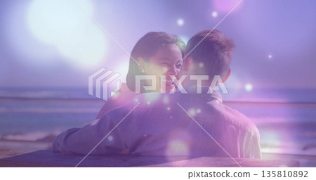 Leaning smiling couple admiring ocean horizon, light flares and bokeh at seaside dusk, with bench 135810892