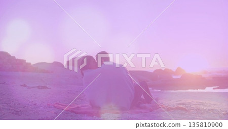 Sitting couple sharing light-colored blanket on pink fringed beach blanket at sunset, pastel light Sitting couple sharing light-colored blanket on pink fringed beach blanket at sunset, pastel light 135810900