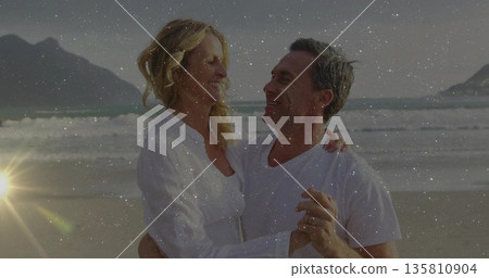 Smiling senior couple embracing near water's edge on sandy beach during sunset, with wedding rings 135810904