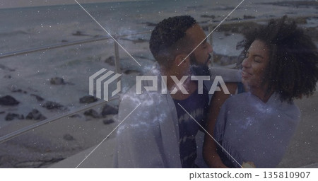 Sitting couple sharing gray blanket by metal railing on seaside promenade, sipping hot drinks Sitting couple sharing gray blanket by metal railing on seaside promenade, sipping hot drinks 135810907