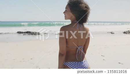 Standing Black woman gazing toward horizon on sandy shoreline with striped bikini, copy space 135811016