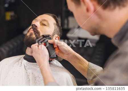 Barber trimming beard of male client with electric clipper in barbershop. Professional grooming process, beard shaping, men haircare routine, classic barber service. Barber trimming beard of male client with electric clipper in barbershop. Professional grooming process, beard shaping, men haircare routine, classic barber service. 135811635