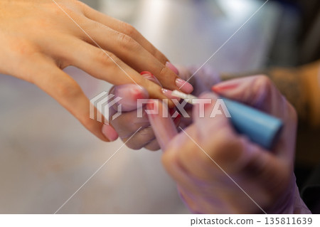 Close up of professional manicure process. Nail technician applying gel polish on female nails with brush while wearing protective gloves in beauty salon Close up of professional manicure process. Nail technician applying gel polish on female nails with brush while wearing protective gloves in beauty salon 135811639