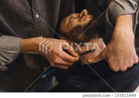 Close up of professional barber shaving male neck with straight razor in barbershop. Beard contouring, classic wet shave, men grooming service and barber work process concept. Close up of professional barber shaving male neck with straight razor in barbershop. Beard contouring, classic wet shave, men grooming service and barber work process concept. 135811649