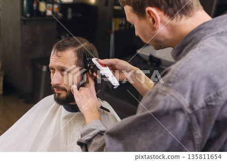 Professional barber trimming client hair with electric clipper in modern barbershop. Male grooming service, precise hairstyling process, focused expression, contemporary salon interior. Professional barber trimming client hair with electric clipper in modern barbershop. Male grooming service, precise hairstyling process, focused expression, contemporary salon interior. 135811654