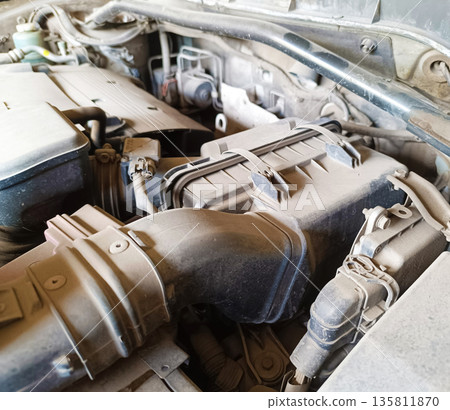 Close-up view of a very dusty and neglected car engine bay, focusing on the air filter housing and various plastic components covered in grime 135811870