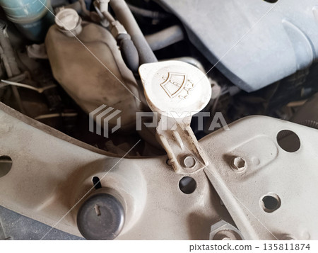 Close-up of a very dusty car engine bay, highlighting the windshield washer fluid reservoir cap with its distinct shield and spray icon. Maintenance needed 135811874