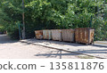 Row of heavily rusted, wheeled industrial dumpsters sits on a dusty patch of ground, backed by lush green trees and a wire fence under bright sunlight. 135811876