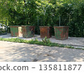 Row of heavily rusted industrial trash bins sits in the bright sun, surrounded by lush green trees and a chain-link fence. Waste disposal 135811877