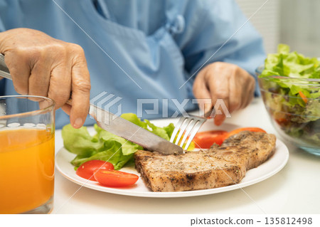 Asian elderly woman patient eating pork chop stake and vegetable salad for healthy food in hospital. 135812498