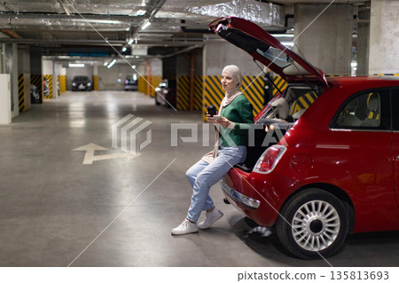 A woman wearing a hijab sits in the open trunk of a red car in an underground parking garage, holding a phone and a coffee cup A woman wearing a hijab sits in the open trunk of a red car in an underground parking garage, holding a phone and a coffee cup 135813693