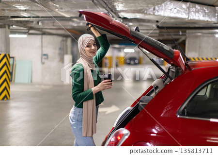 A woman wearing a hijab smiles while opening the trunk of her red car in a parking garage A woman wearing a hijab smiles while opening the trunk of her red car in a parking garage 135813711