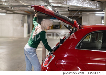 A woman wearing a hijab loads the trunk of a red car in an underground parking garage A woman wearing a hijab loads the trunk of a red car in an underground parking garage 135813713